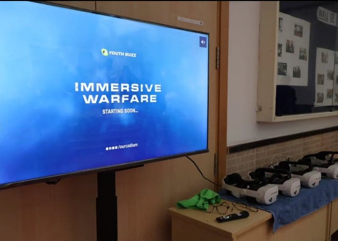 The session setup — a large display reading Immersive Warfare beside a row of charged VR headsets ready for students